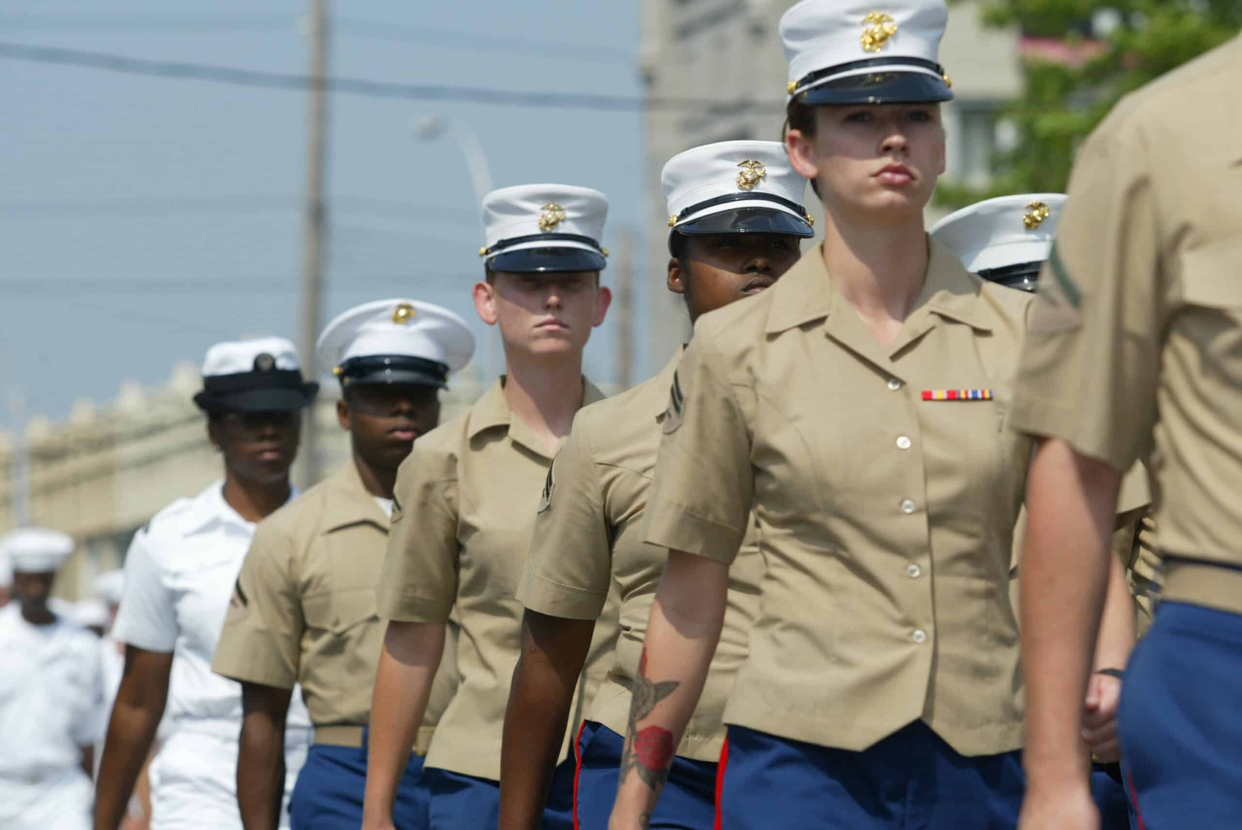 A group of uniformed Marines march in formation outdoors, with several Navy personnel visible in the background.